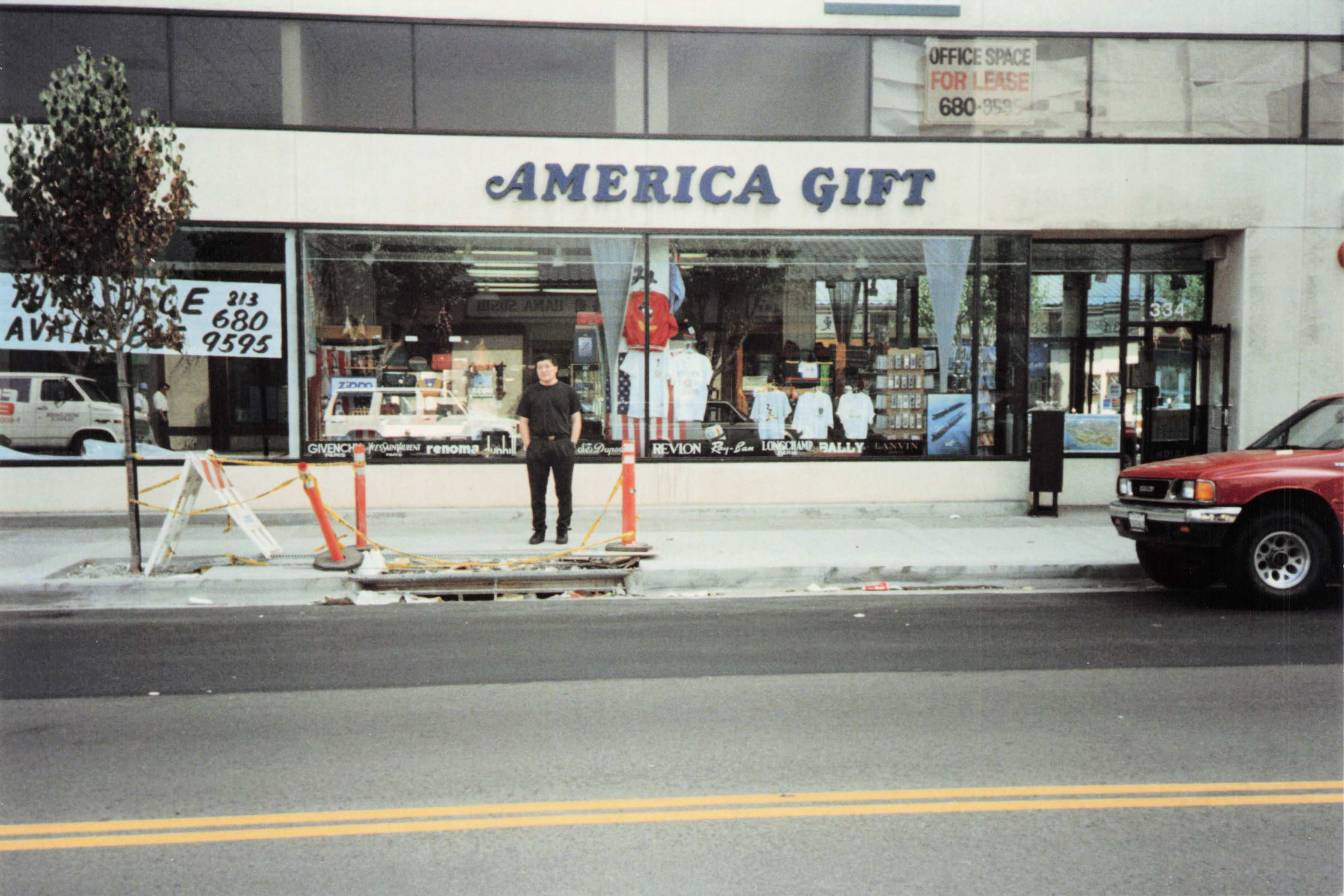 Minsoo Kang in all-black, deadpan, in front of the AMERICA GIFT store on an LA street