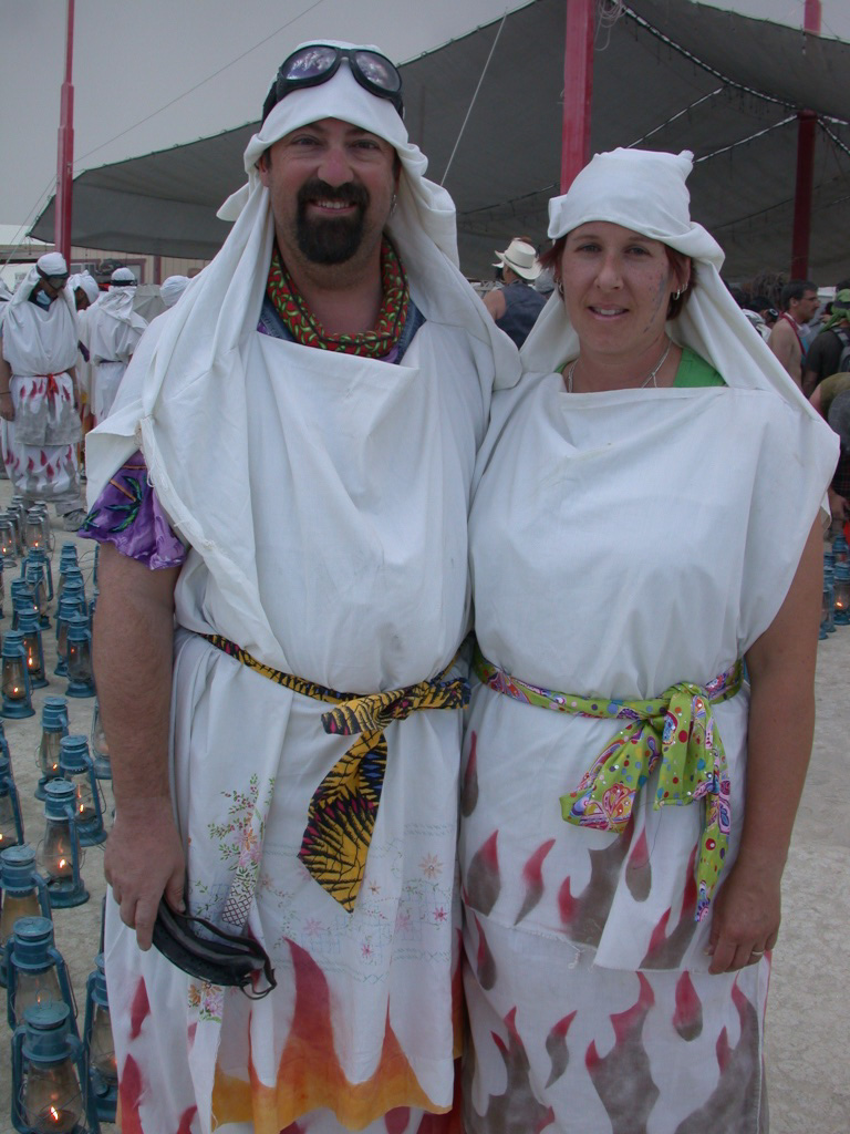 Fred and Becke in matching white flame-hemmed robes amid rows of lit blue hurricane lanterns — Burning Man Lamplighters, 2007.