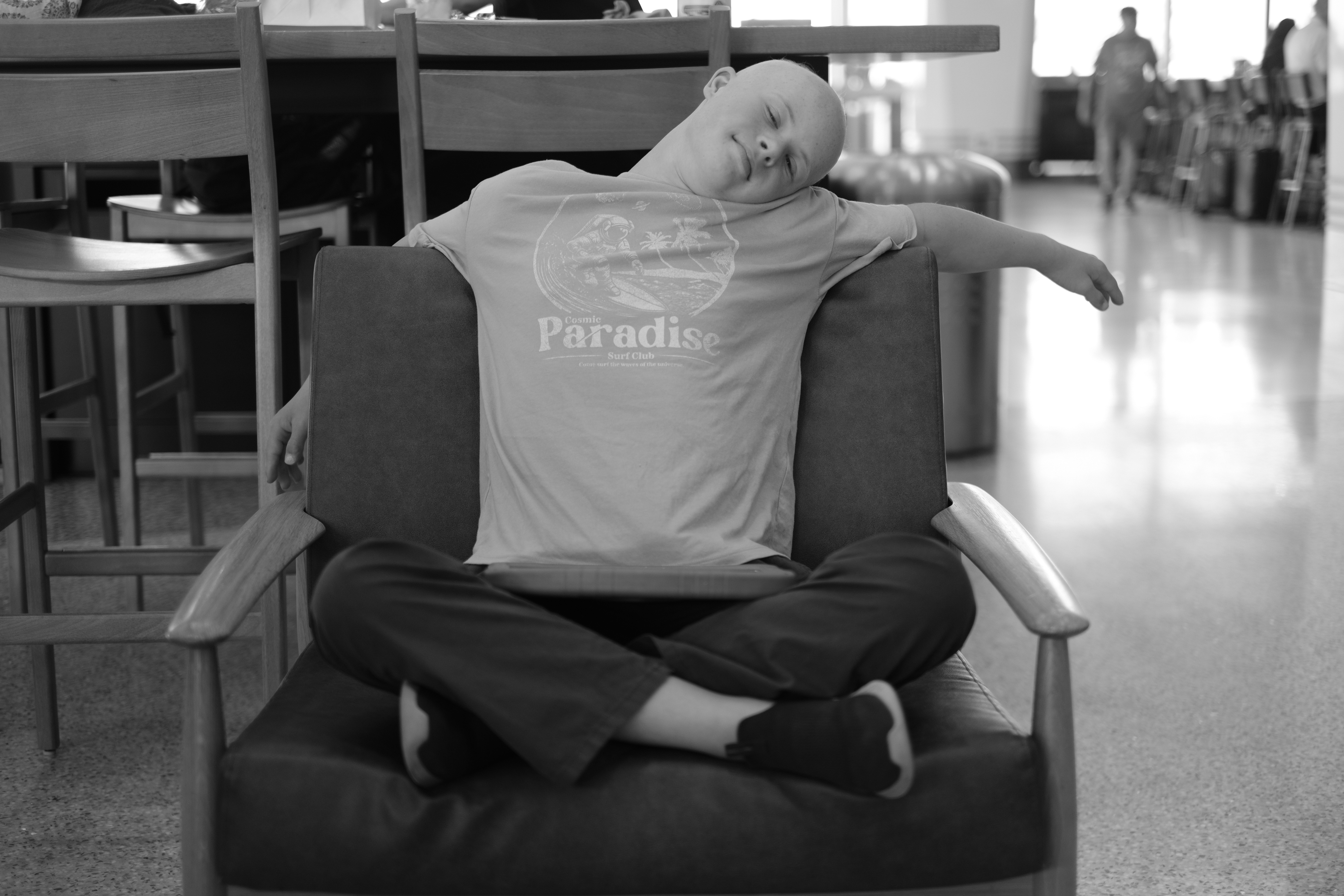 Black-and-white photo of Dylan sprawled sideways across an airport lounge chair, one arm draped over the back, other travelers and luggage in the background.