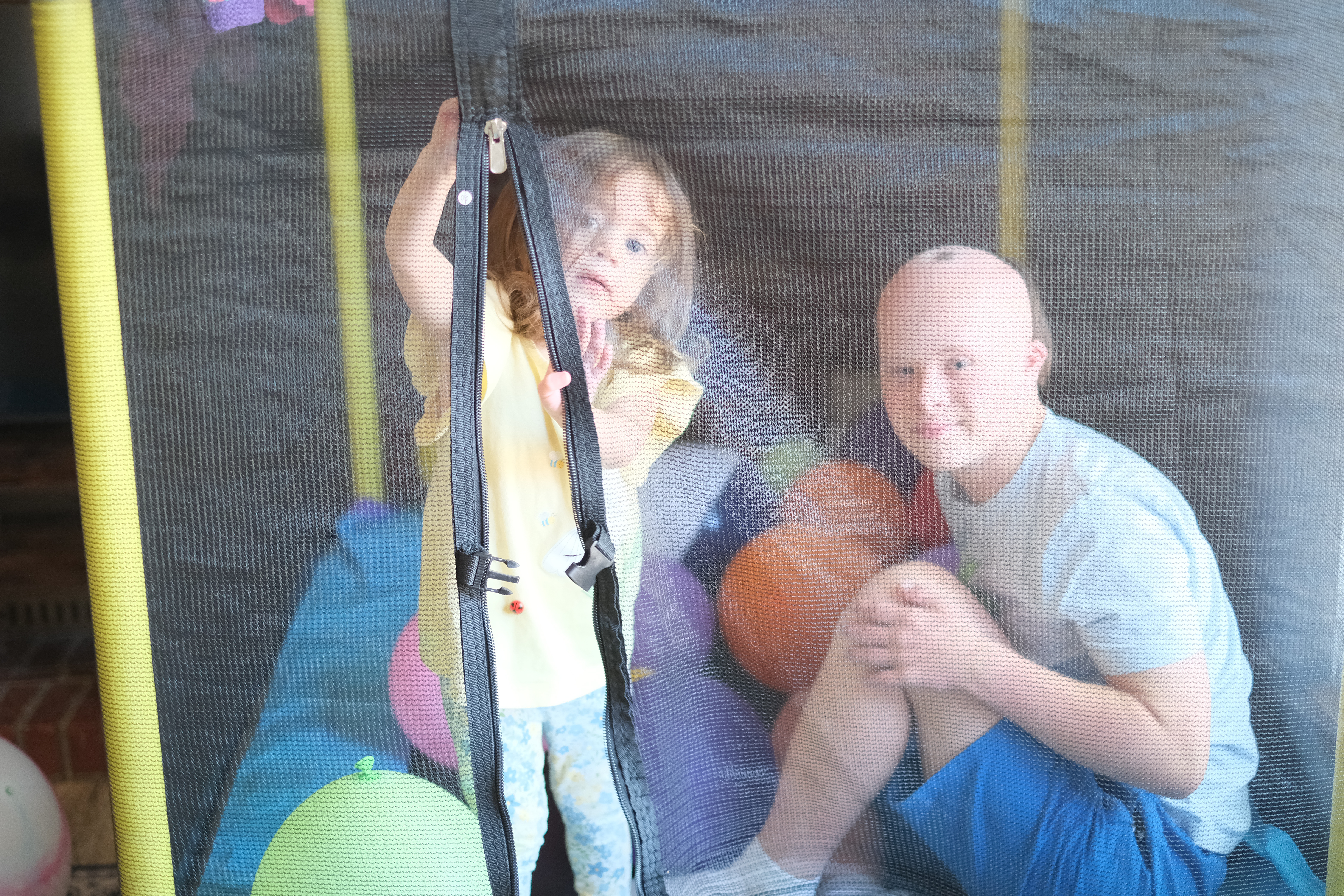 Photo taken through the mesh wall of a pop-up ball-pit tent, showing Dylan and a young child crouched together among the colorful balls.