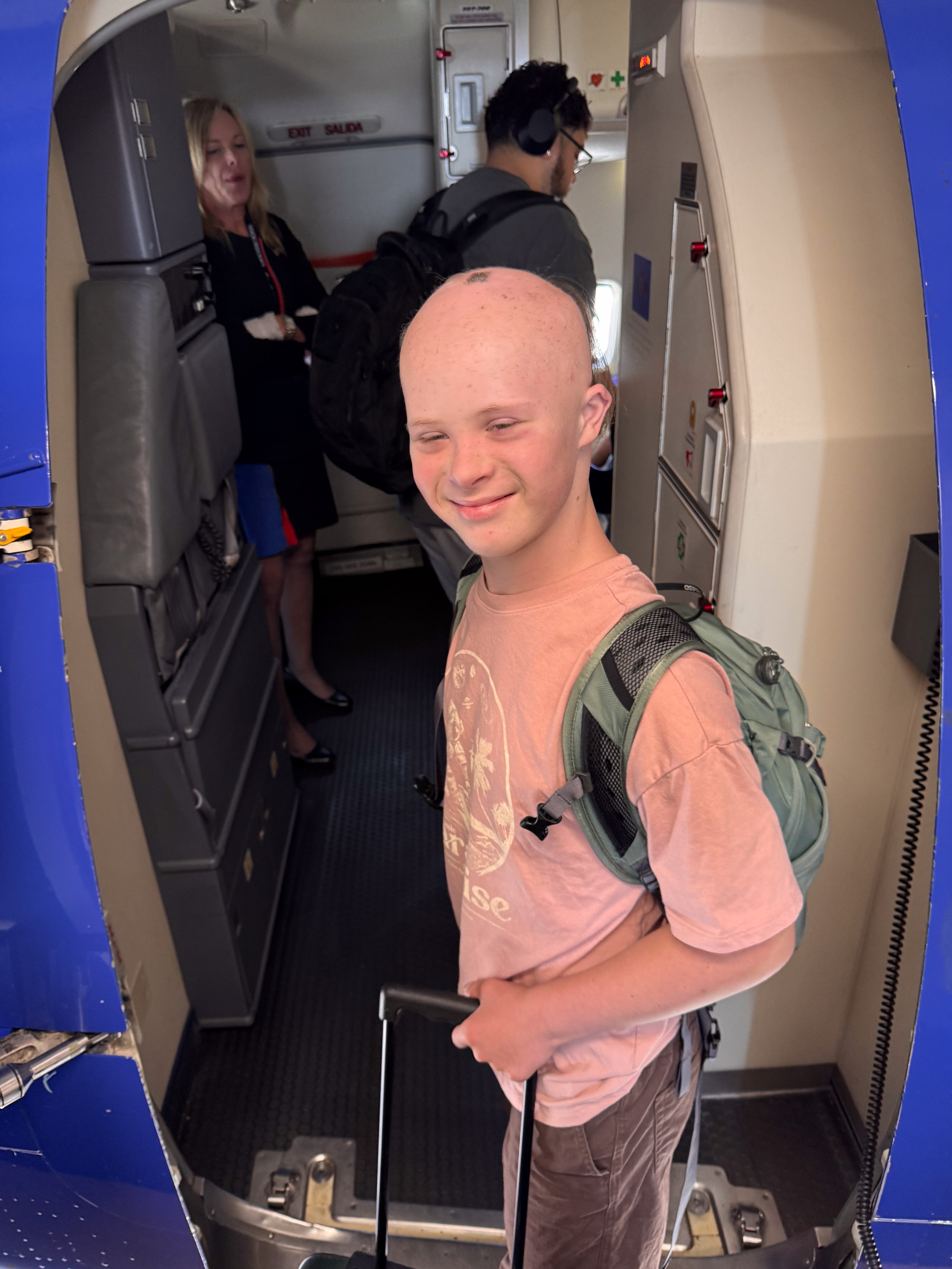 Dylan boarding the plane at the jet bridge, backpack and rolling suitcase, a flight attendant visible in the cabin behind him.