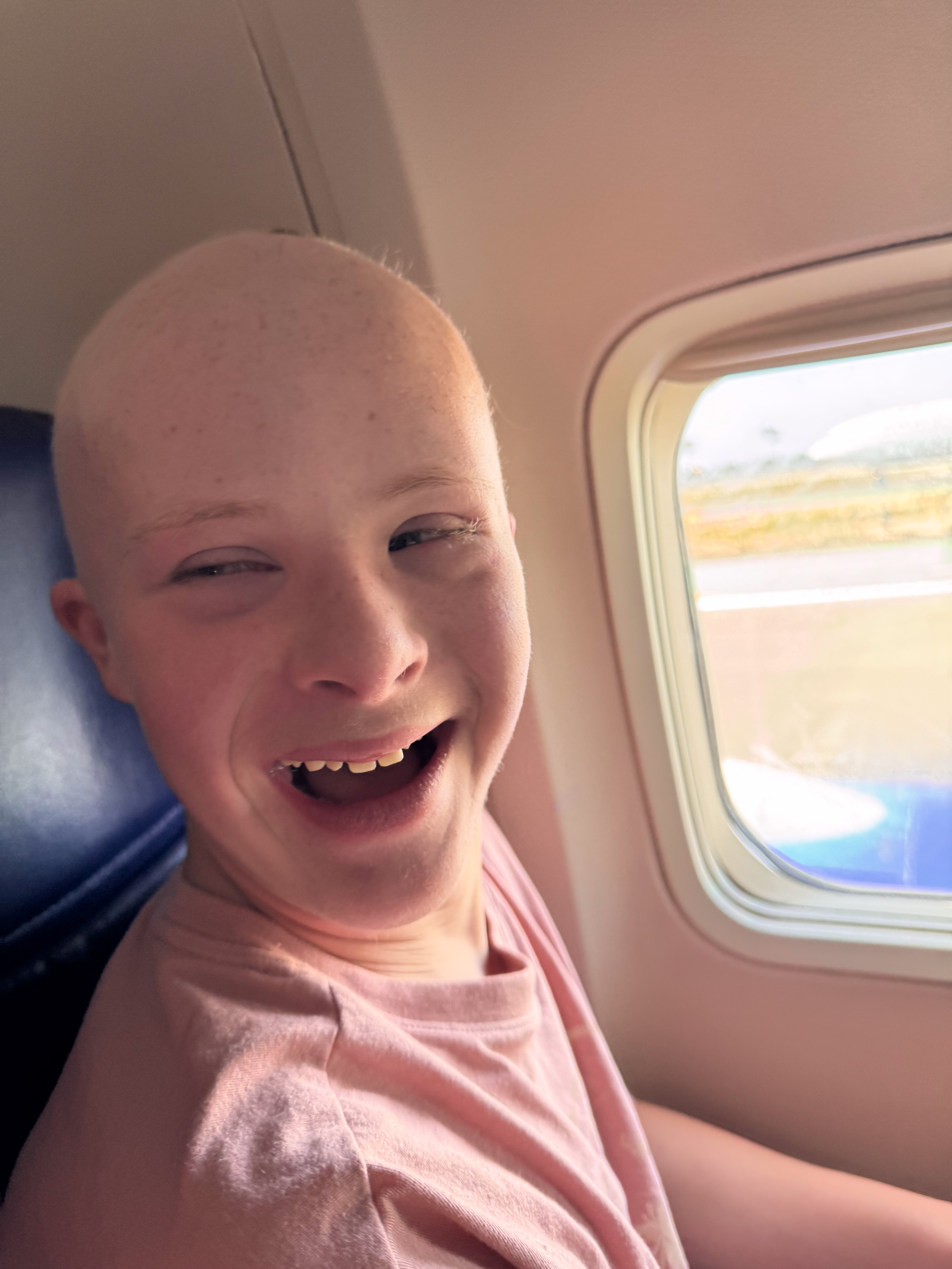 Dylan laughing in his airplane window seat, tarmac and another plane visible outside.
