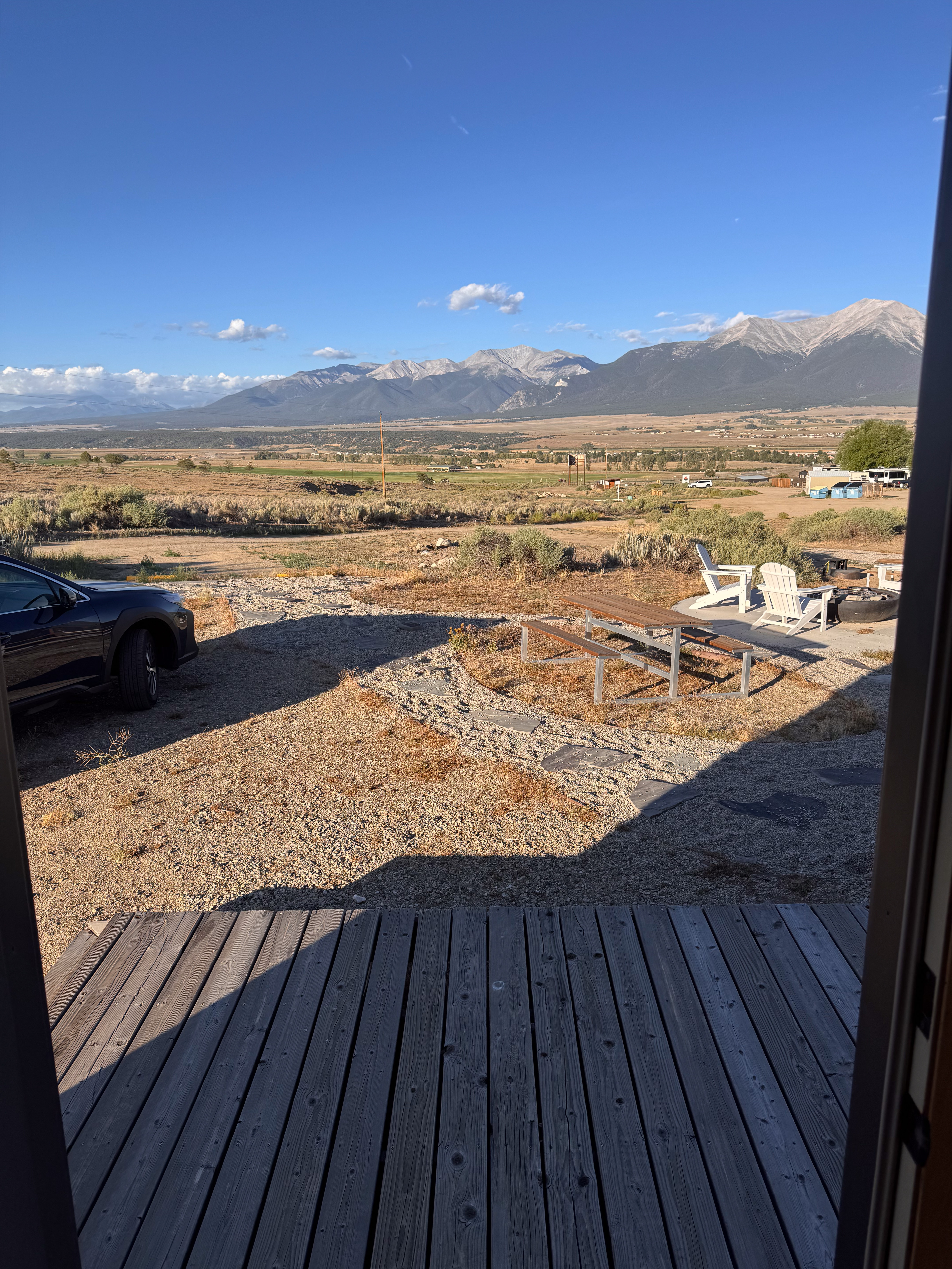 View from a lodging deck: picnic table, Adirondack chairs, a fire pit, and a broad valley with snow-streaked peaks.