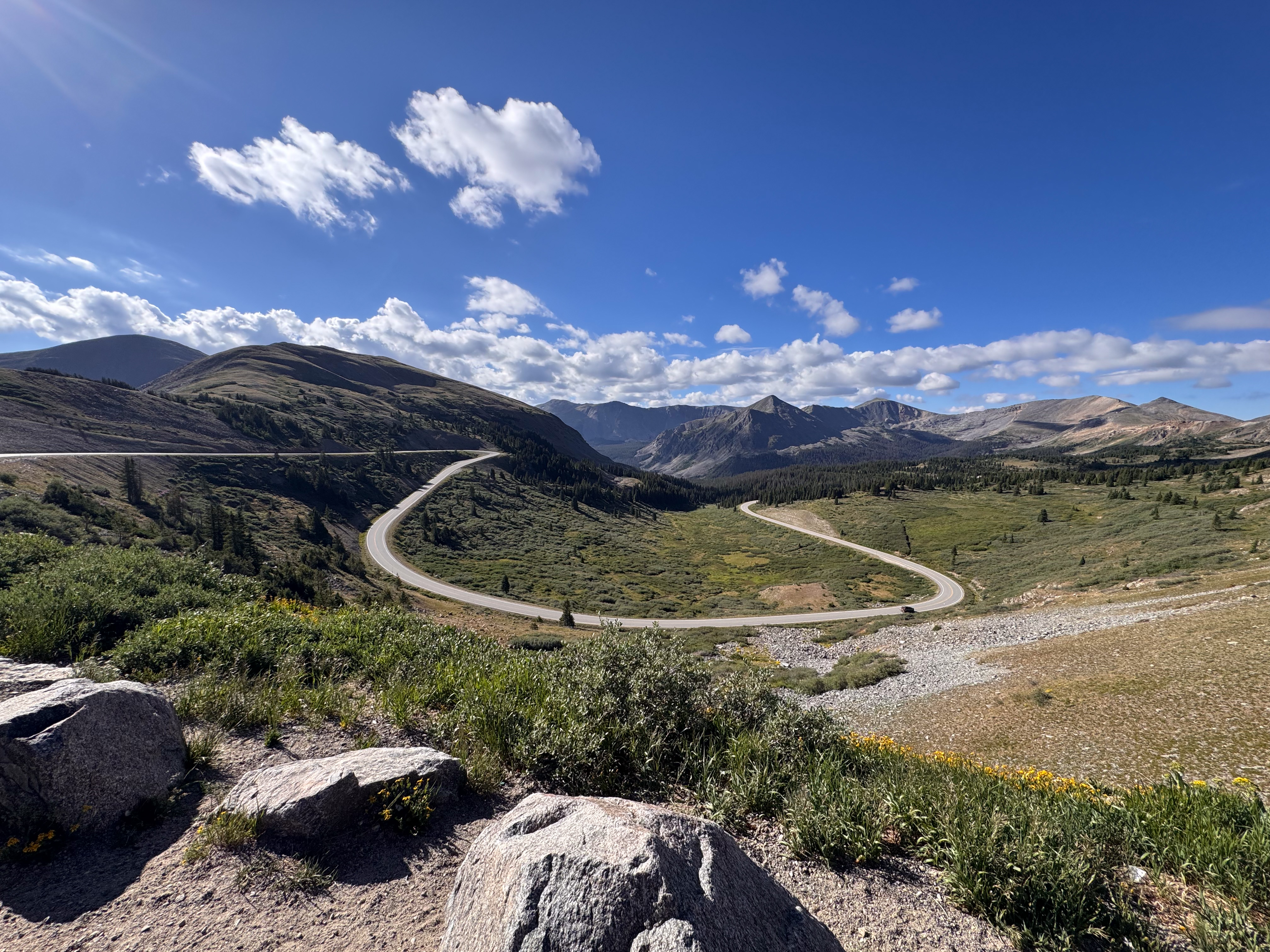 A sweeping S-curve of paved mountain highway through an alpine valley, granite boulders in the foreground, peaks and clouds beyond.