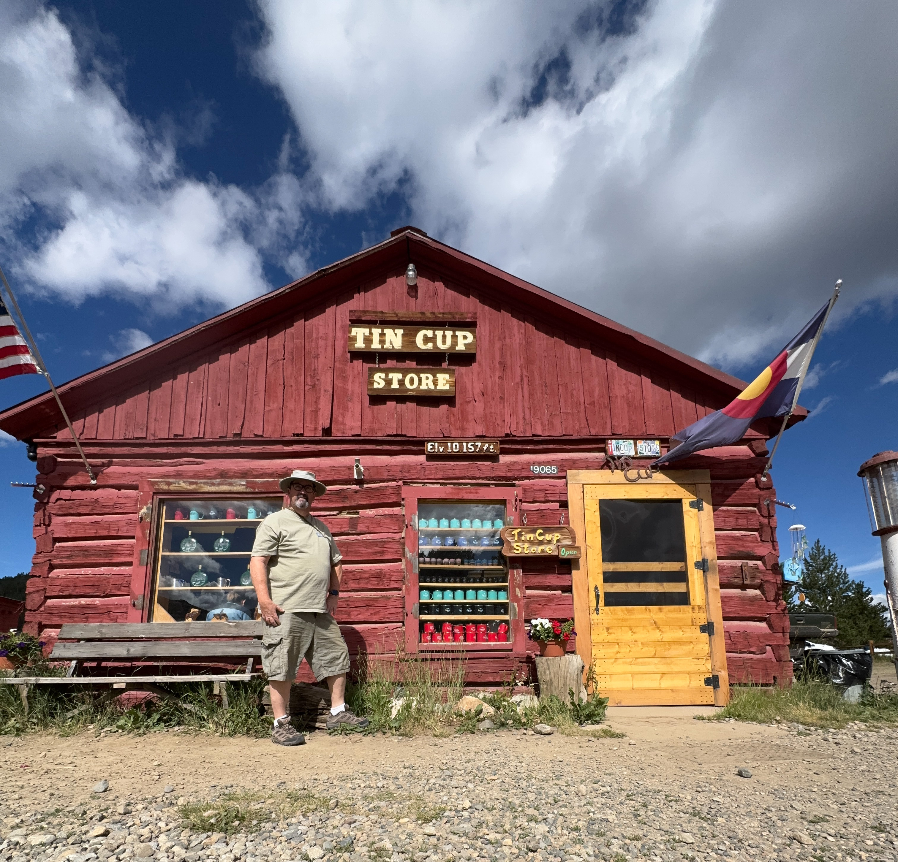Fred standing beside the Tin Cup Store, a log cabin with a sign reading "Elv 10,157 ft" and a Colorado state flag flying.