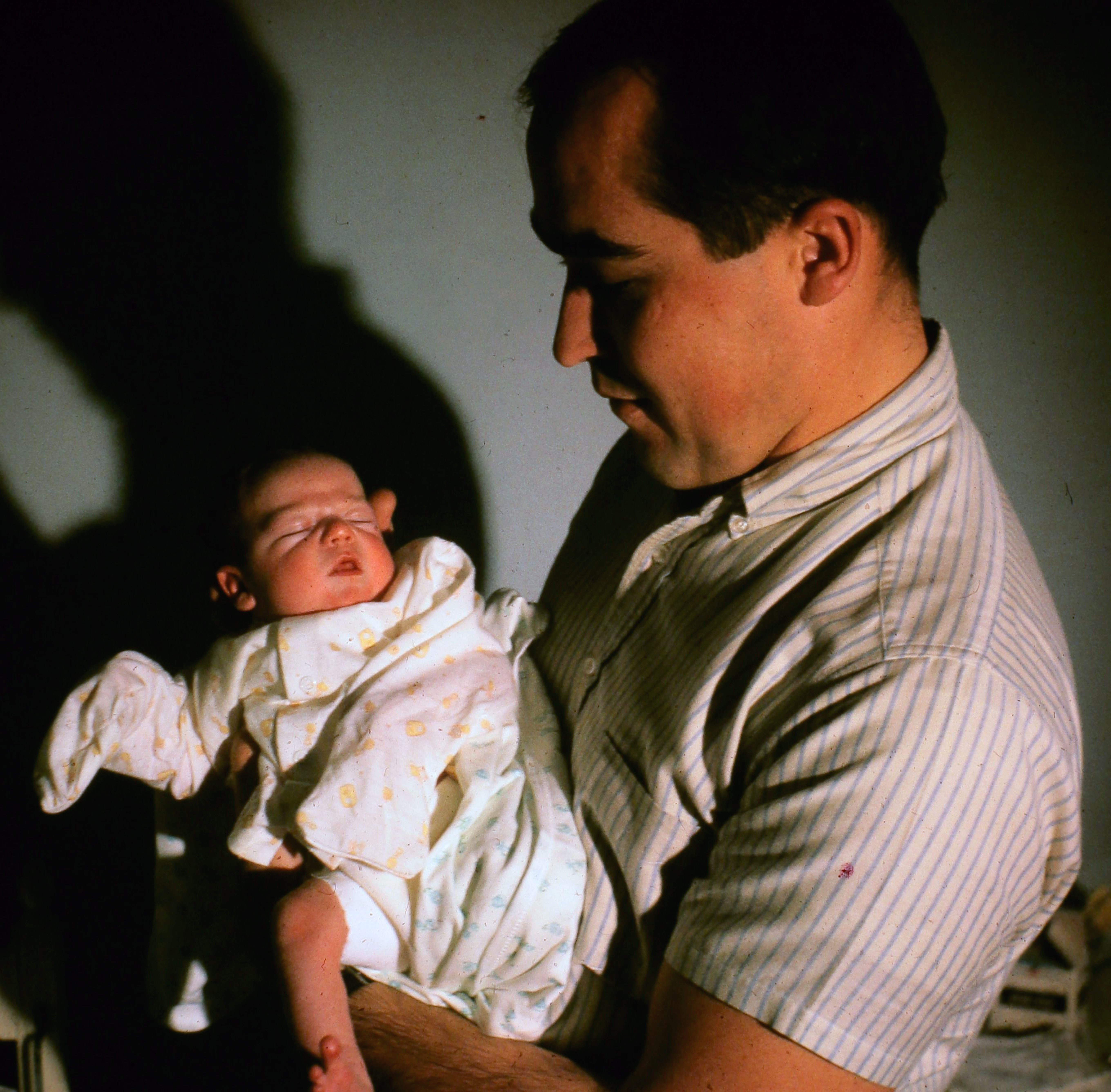 Phil, young, holding his swaddled firstborn under dramatic side-light — El Centro, est. late 1965 or early 1966.