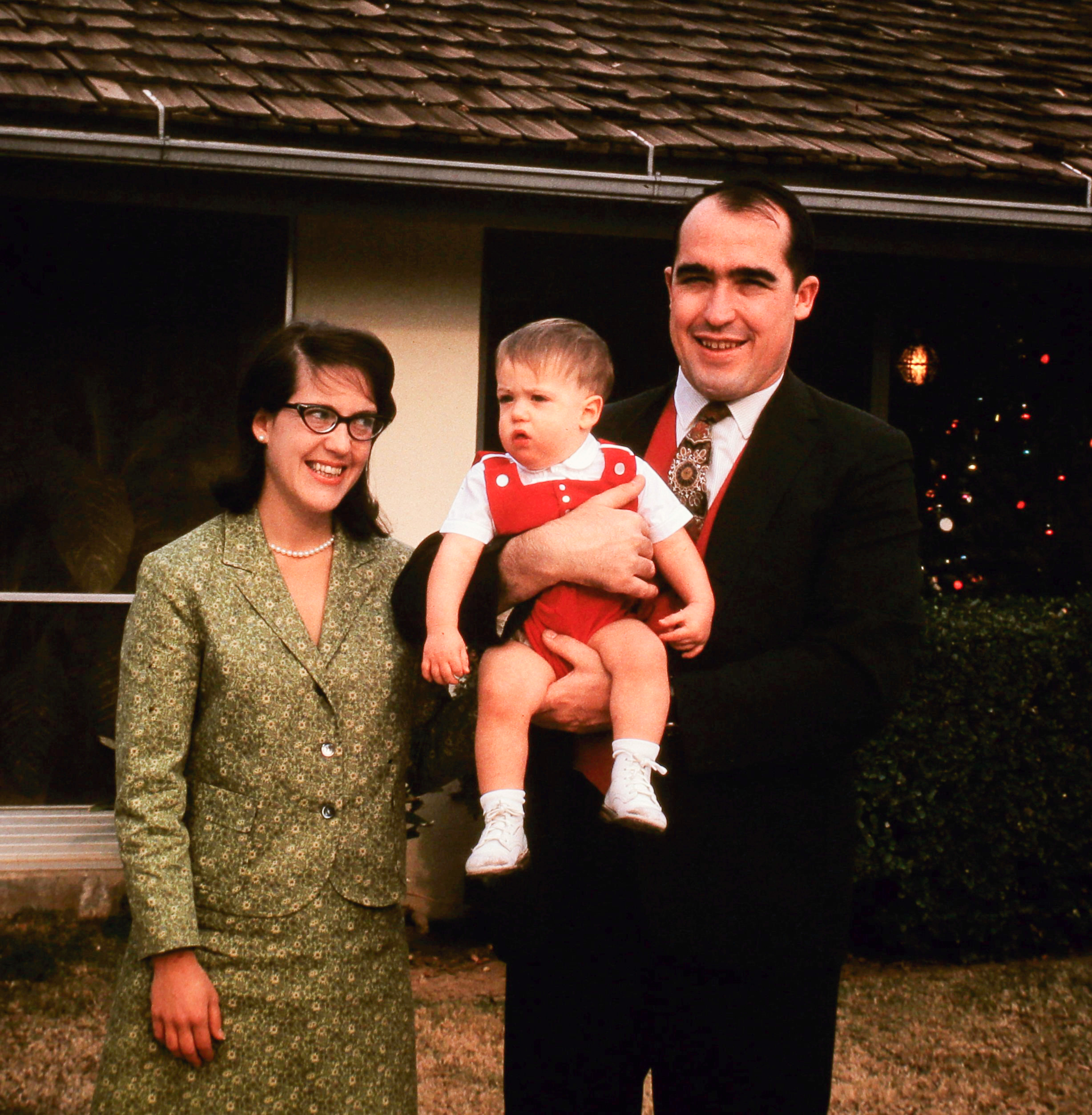 Phil, Elise, and baby Fred outside the house — Elise in cat-eye glasses and green tweed, Fred in red overalls. El Centro, Christmas 1966.