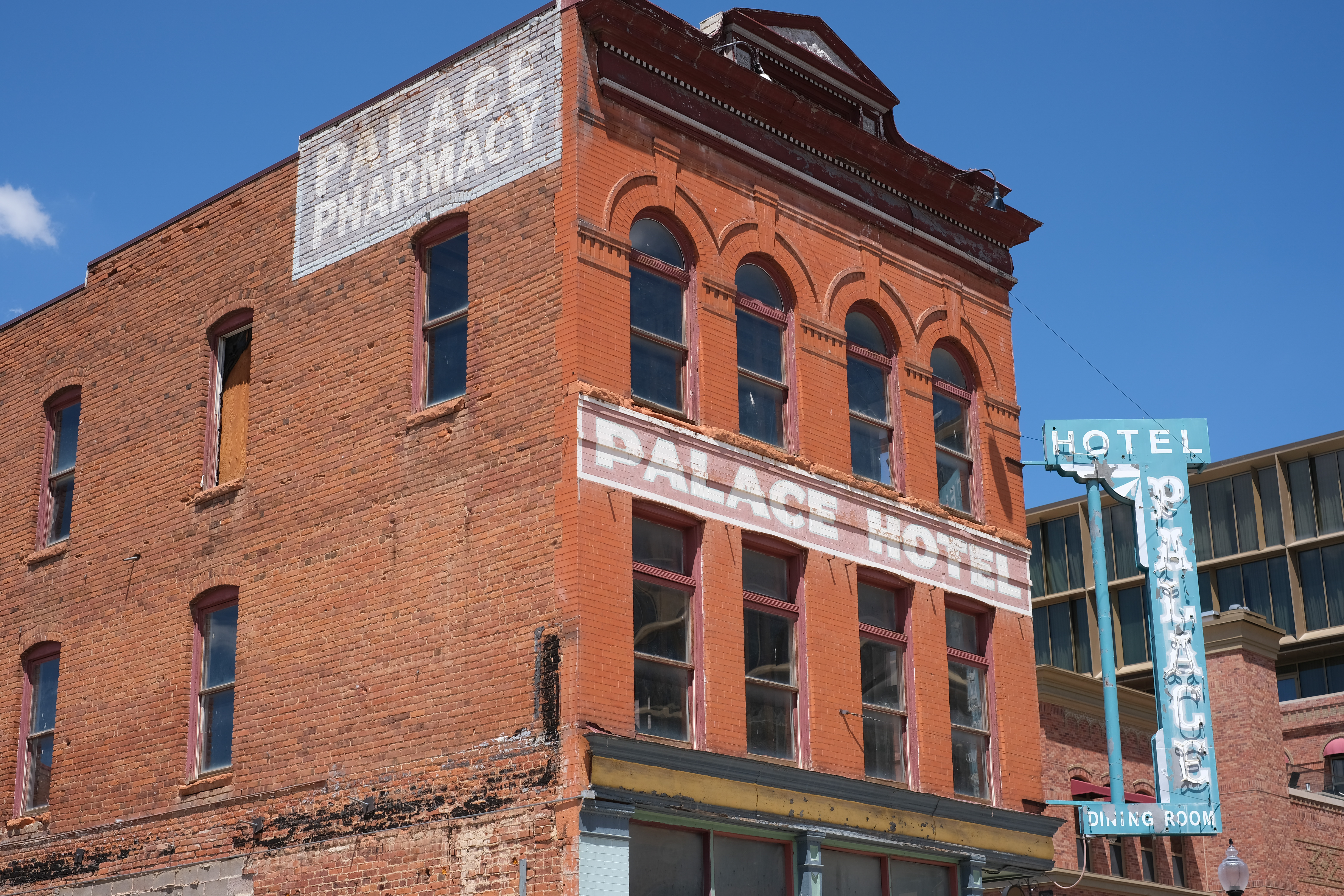 Corner of a brick building against blue sky — faded painted signs read "PALACE PHARMACY" and "PALACE HOTEL," with a vintage teal neon "HOTEL / PALACE / DINING ROOM" sign projecting from the corner.