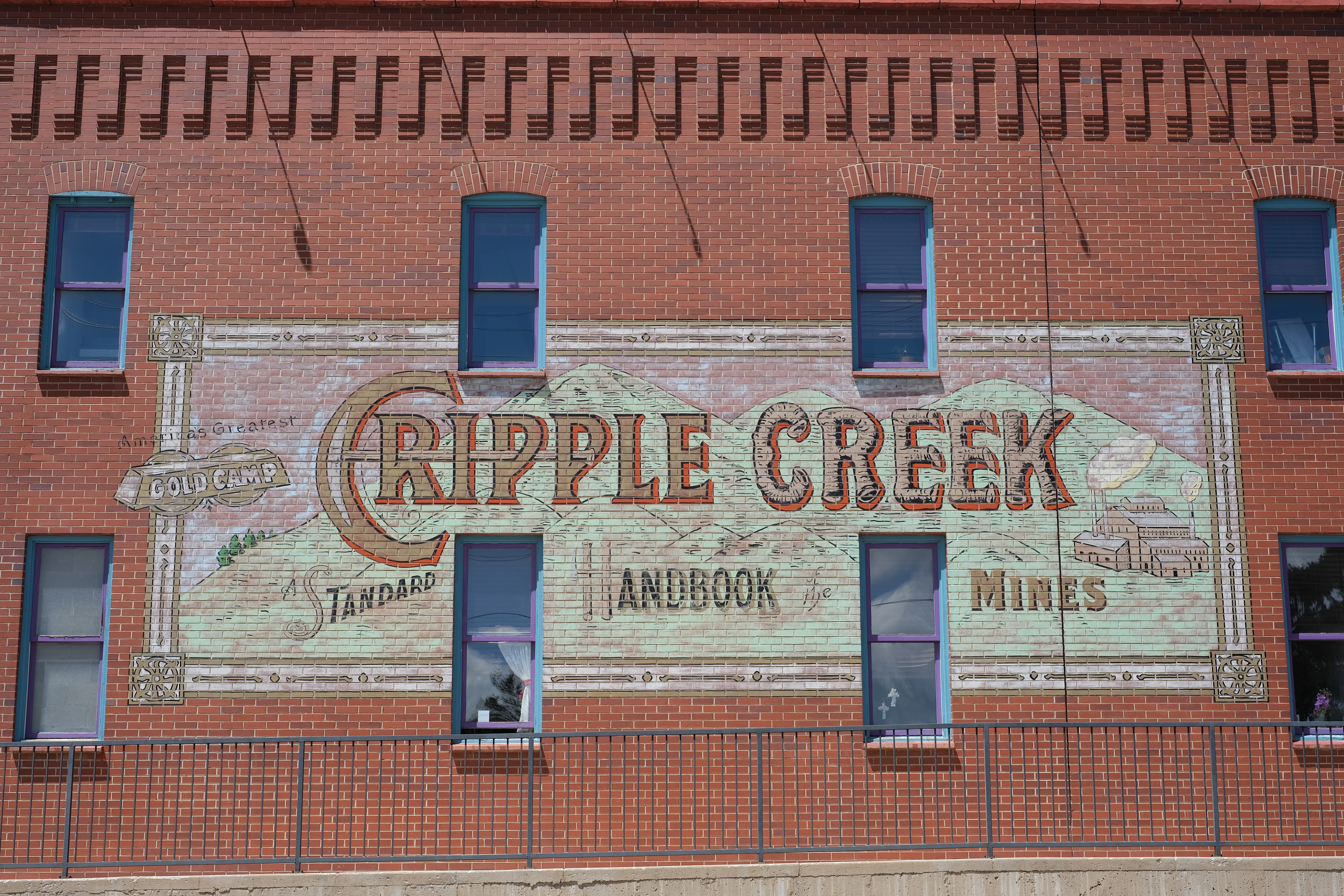 A large hand-painted ghost sign spanning a brick facade in Cripple Creek: "CRIPPLE CREEK" in bold letters over a painted mountain range, with "America's Greatest Gold Camp" and a small mine/smokestack graphic.