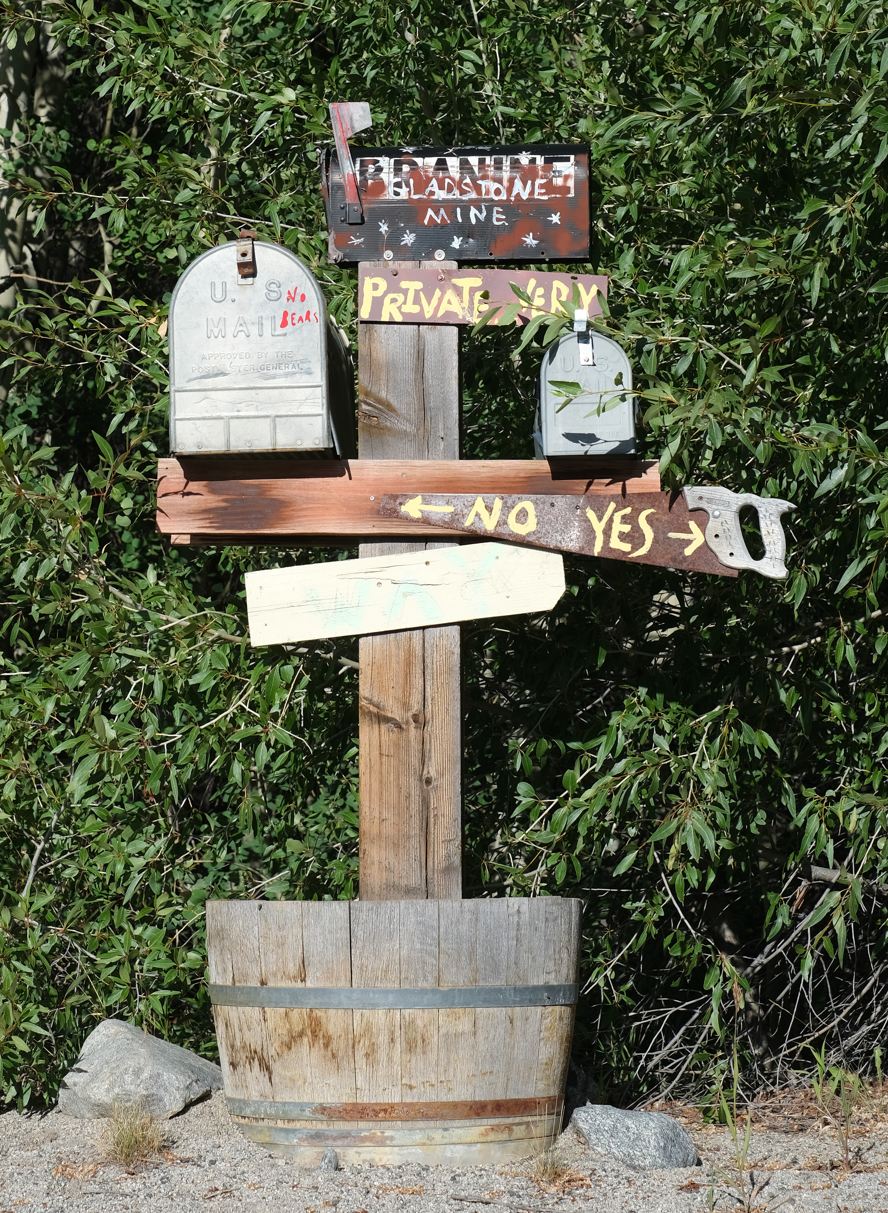A folk-art roadside signpost — hand-painted "Gladstone Mine" sign, mismatched mailboxes (one warning "NO BEARS"), and a rusty hand-saw repurposed as a directional sign, mounted in an old wine barrel.