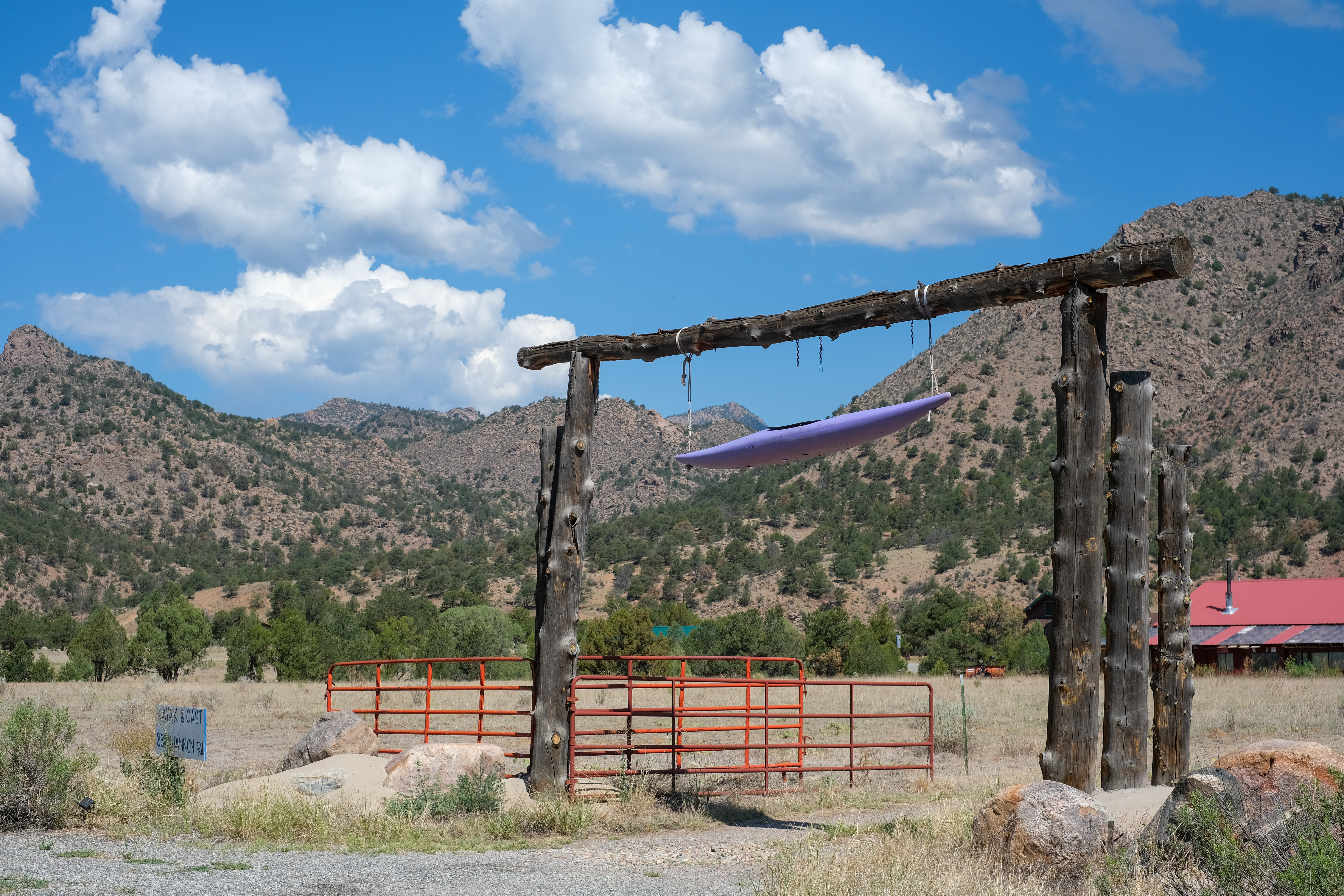 A log-post ranch gate strung with an orange metal cattle gate, a kayak hung from the crossbeam, and a hand-lettered "Kayak & Cast" sign on a boulder.