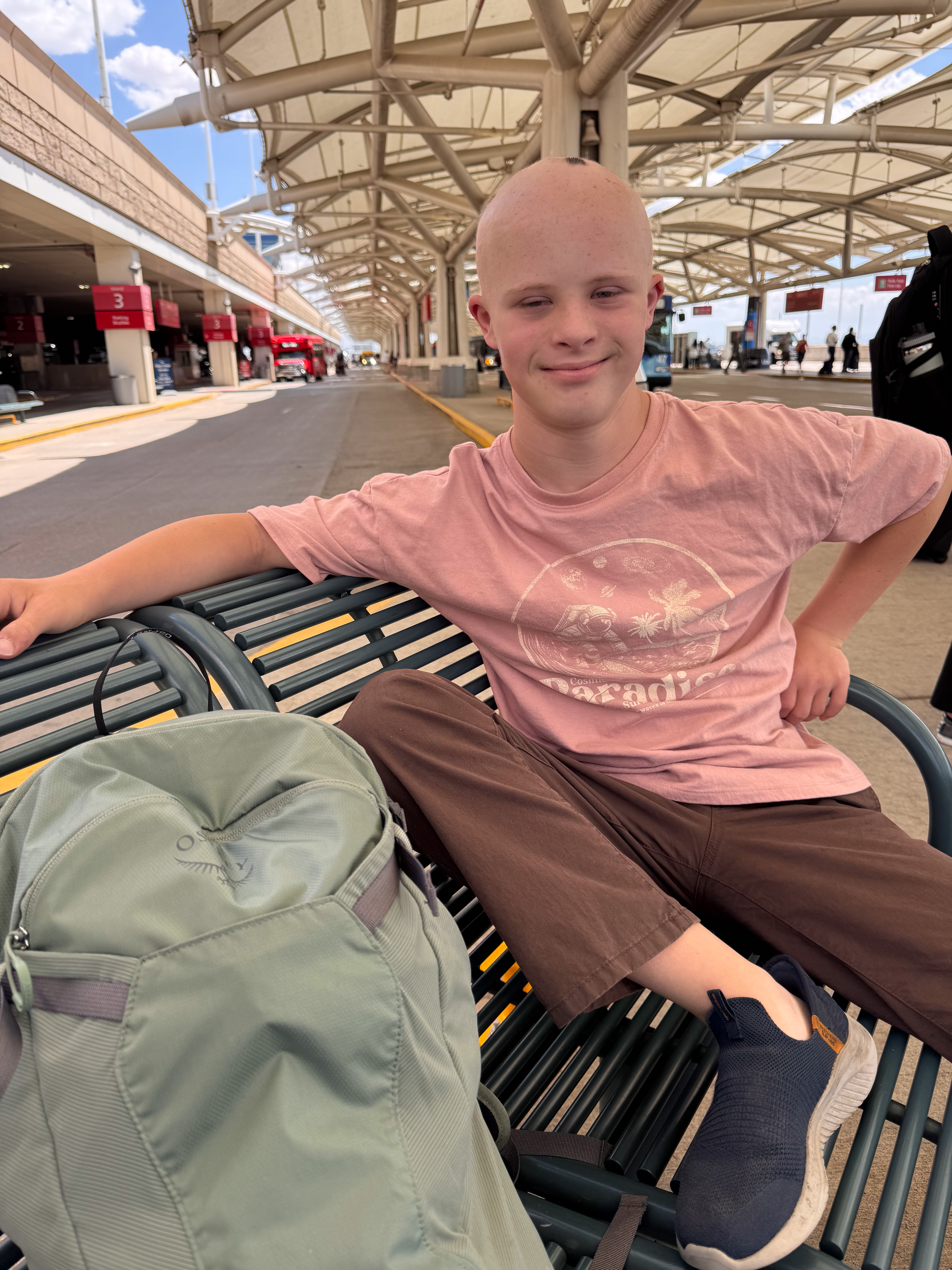 Dylan sitting on a bench under a large tension-fabric canopy at the airport, buses and numbered gate signage behind him.