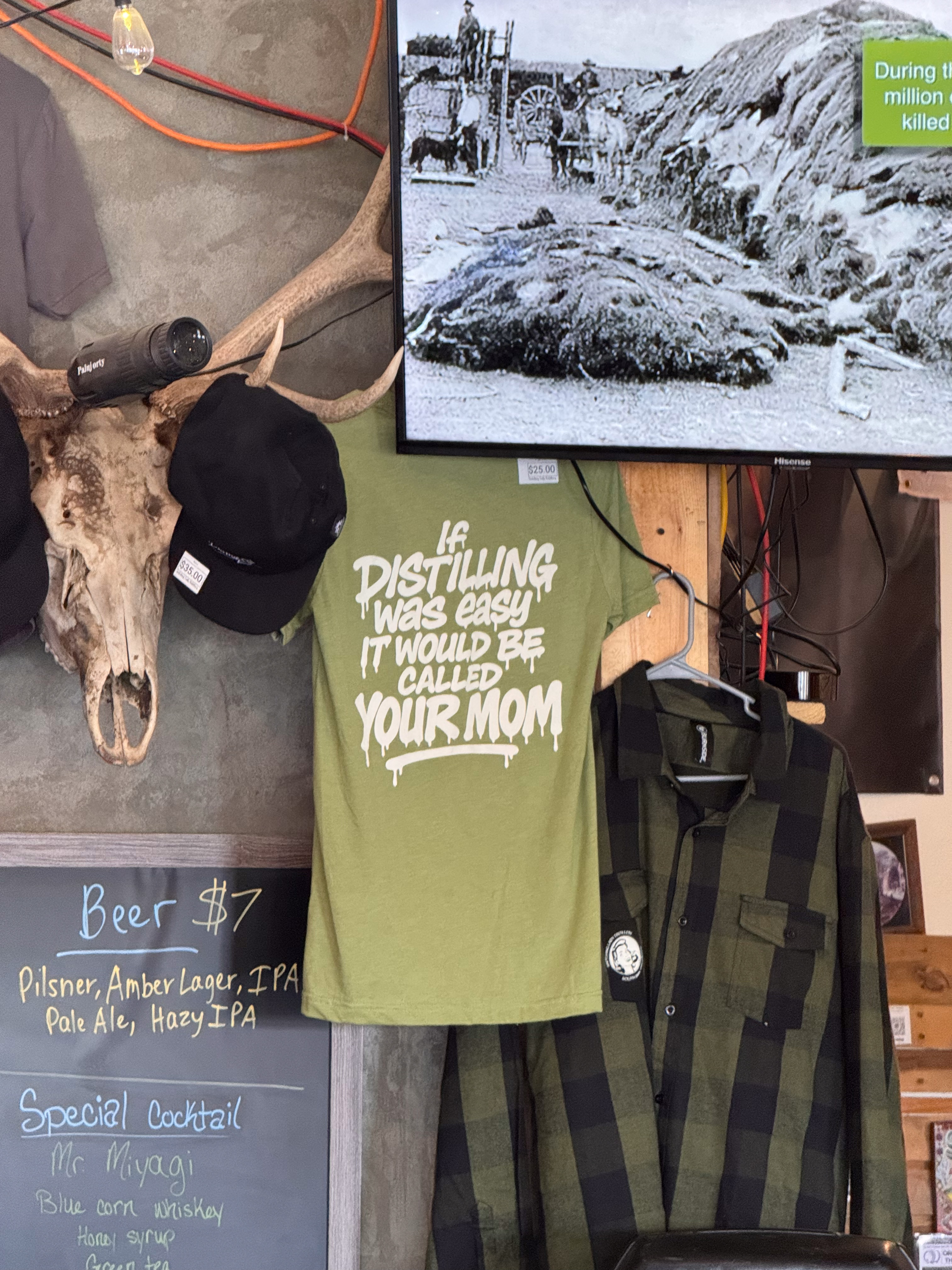Wall of a distillery — antler mounts, a novelty T-shirt ("If distilling was easy..."), and an old buffalo-hunt photo playing on the TV.