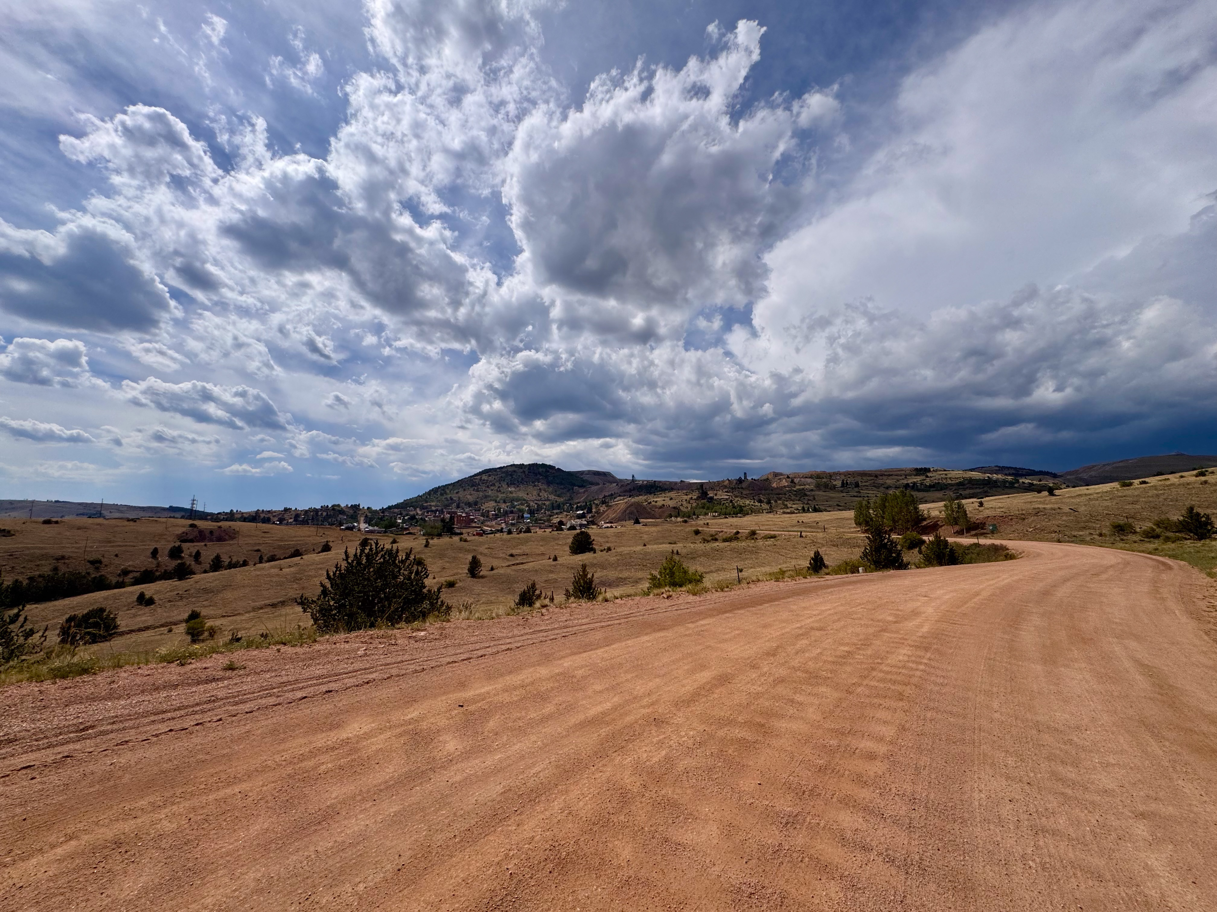 A red dirt road descending into a valley, with a small mining town and mine headframe visible on the hillside under a storm-lit sky.